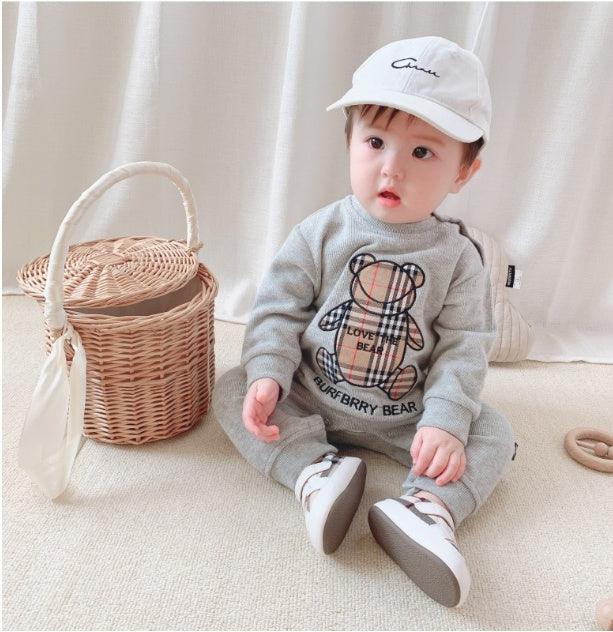 Baby in grey knit bear-print sweatshirt and pants, white cap, and sneakers, sitting indoors