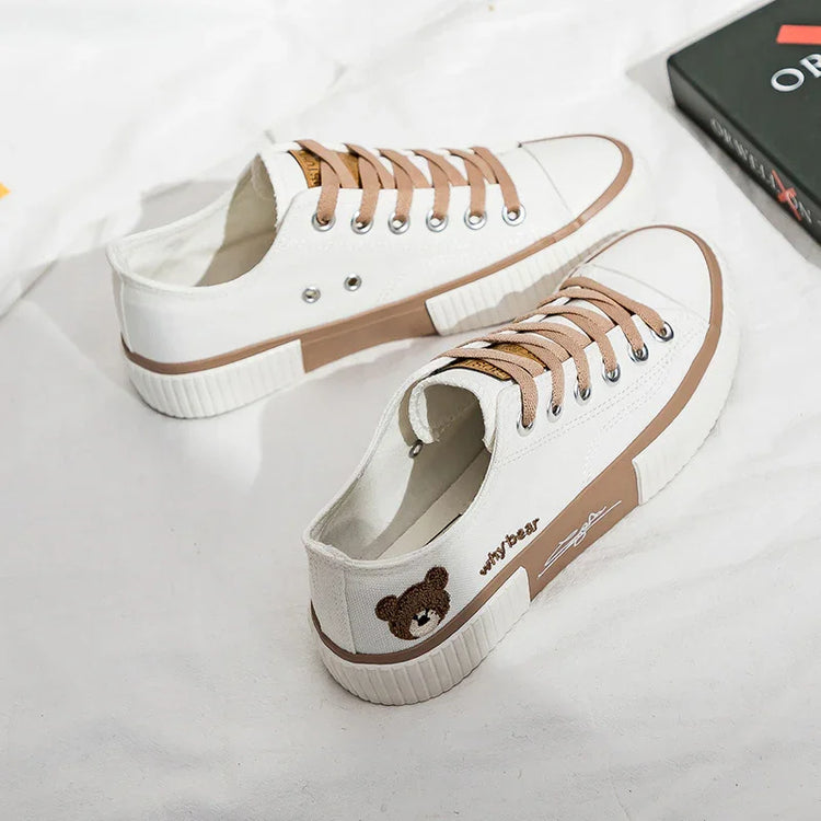White canvas low-top sneakers with tan accents, flat laces, and embroidered bear detail.