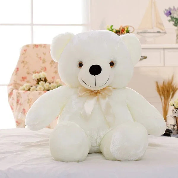 Plush white teddy bear with a ribbon bow, soft fabric, sitting on a bed in a cozy room.