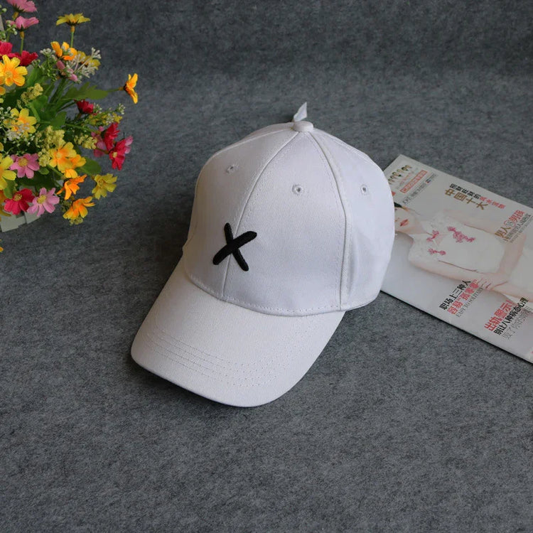 White cotton baseball cap with black embroidered X, adjustable fit, on grey surface.