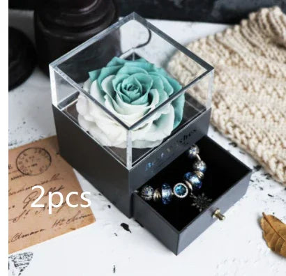 Aqua and white preserved rose in clear box with black jewelry drawer, decorative bracelet inside.