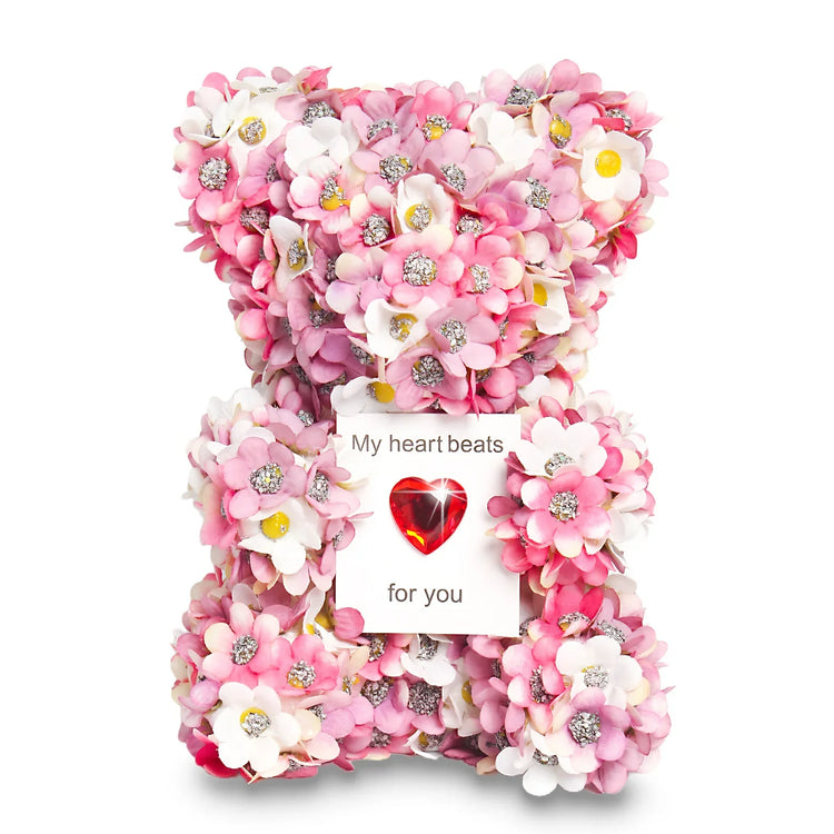 Decorative teddy bear covered in pink and white artificial flowers, holding a heart-shaped card.