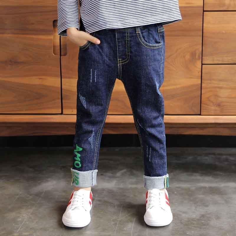 Child wearing relaxed fit dark blue denim jeans with rolled cuffs and embroidered detail, paired with striped shirt and white sneakers.