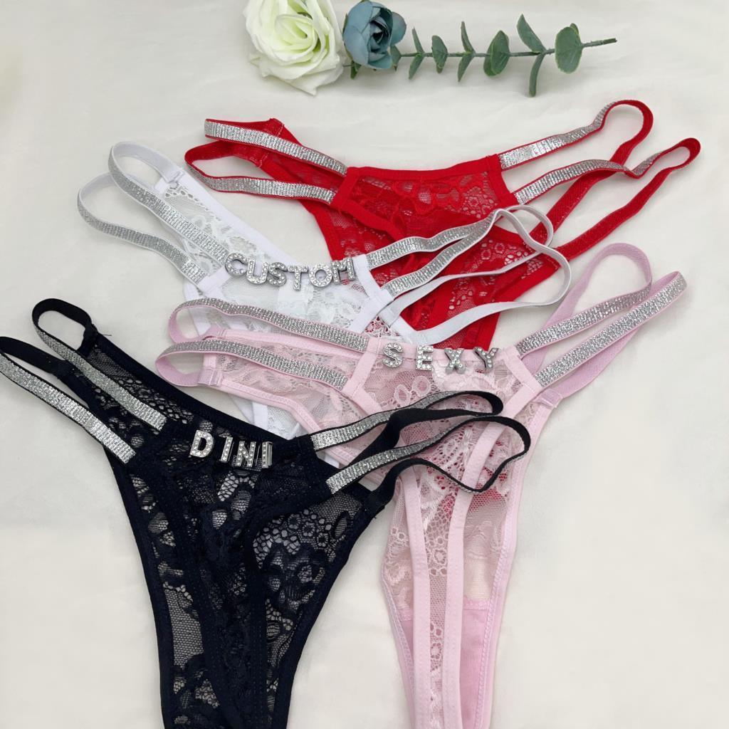Assorted lace thong underwear in black, red, white and pink with rhinestone strap details and custom lettering, displayed on a neutral surface.