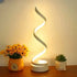Modern spiral LED table lamp with warm light on wooden surface, flowers and succulent beside