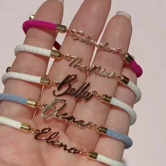 Hand holding colorful cord bracelets with gold nameplate charms, featuring white, pink, and blue bands.
