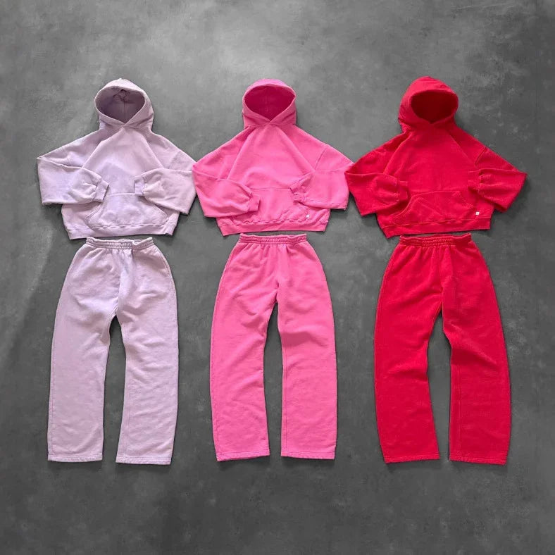 Three sets of cropped hooded sweatshirts and matching jogger pants in pastel purple, bright pink, and red fleece, laid flat on a gray background, showcasing relaxed streetwear style and loose fit.
