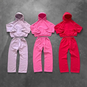 Three sets of cropped hooded sweatshirts and matching jogger pants in pastel purple, bright pink, and red fleece, laid flat on a gray background, showcasing relaxed streetwear style and loose fit.