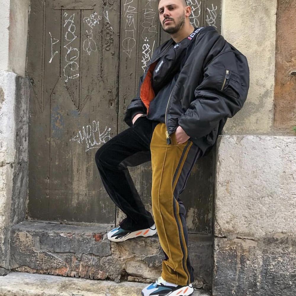 Man in oversized black bomber jacket, velvet mustard and black track pants, and chunky sneakers.