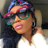 Woman wearing oversized square sunglasses with red and green glitter frame, blue printed headscarf, white faux fur coat, and hoop earrings.