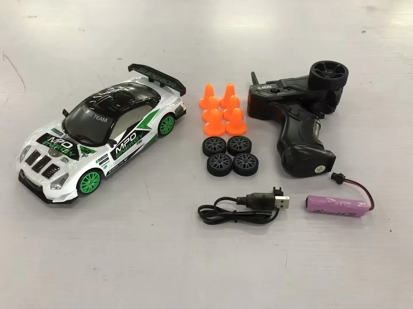Remote control race car set with white and green sport car, black controller, orange cones, spare wheels, charger, and battery on light background.