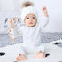 Infant in white long-sleeve cotton romper and knit hat with faux fur pompom, sitting on a bed