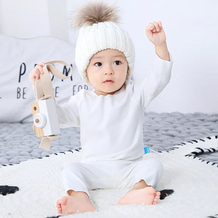 Infant in white long-sleeve cotton romper and knit hat with faux fur pompom, sitting on a bed