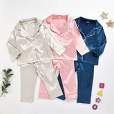 Satin pajama sets for kids in beige, pink, and navy blue with long sleeves and button-up tops.