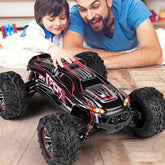 Smiling child in a white shirt playing with large black and red remote control truck indoors.