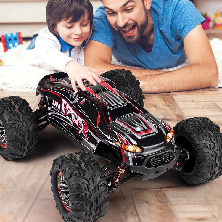 Smiling child in a white shirt playing with large black and red remote control truck indoors.