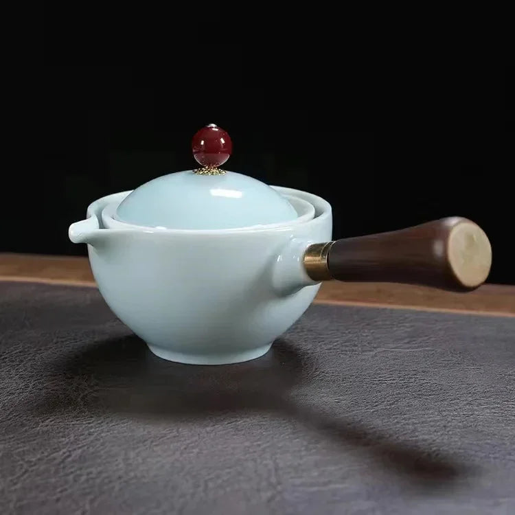 Ceramic teapot with wooden handle, light blue glaze, and round red lid knob on table