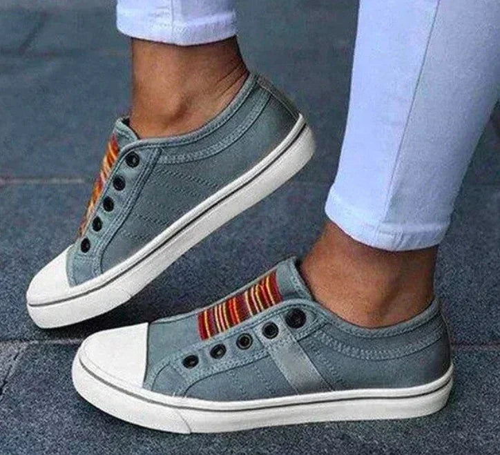 Gray slip-on canvas sneakers with colorful striped elastic and white soles, worn with white pants
