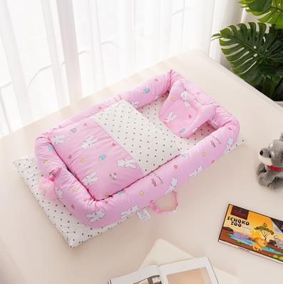 Pink cotton baby lounger with bunny print, soft padded sides, and matching pillow on a bed.
