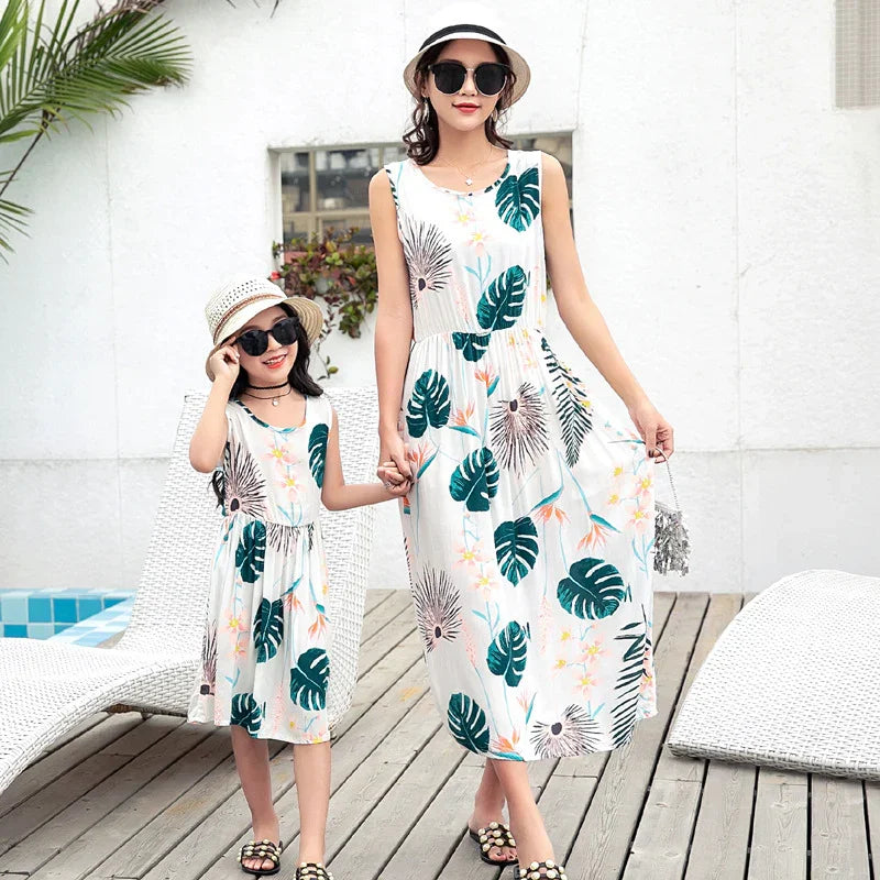Mother and daughter wear matching sleeveless white sundresses with tropical leaf print, relaxed fit.