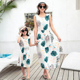 Mother and daughter wear matching sleeveless white sundresses with tropical leaf print, relaxed fit.