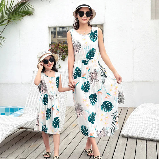 Mother and daughter wear matching sleeveless white sundresses with tropical leaf print, relaxed fit.