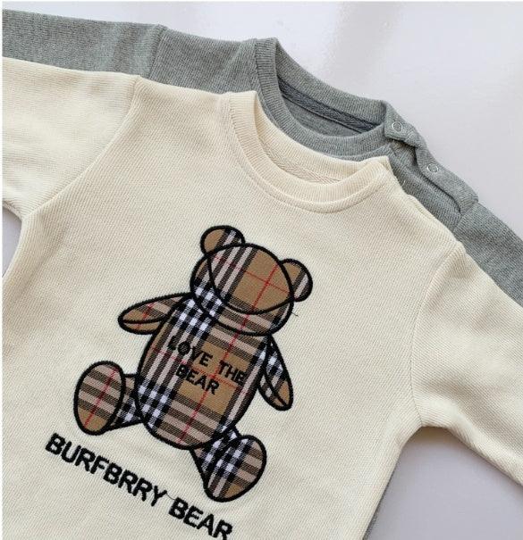 Cream ribbed knit baby romper with plaid teddy bear graphic and text, layered over a gray romper