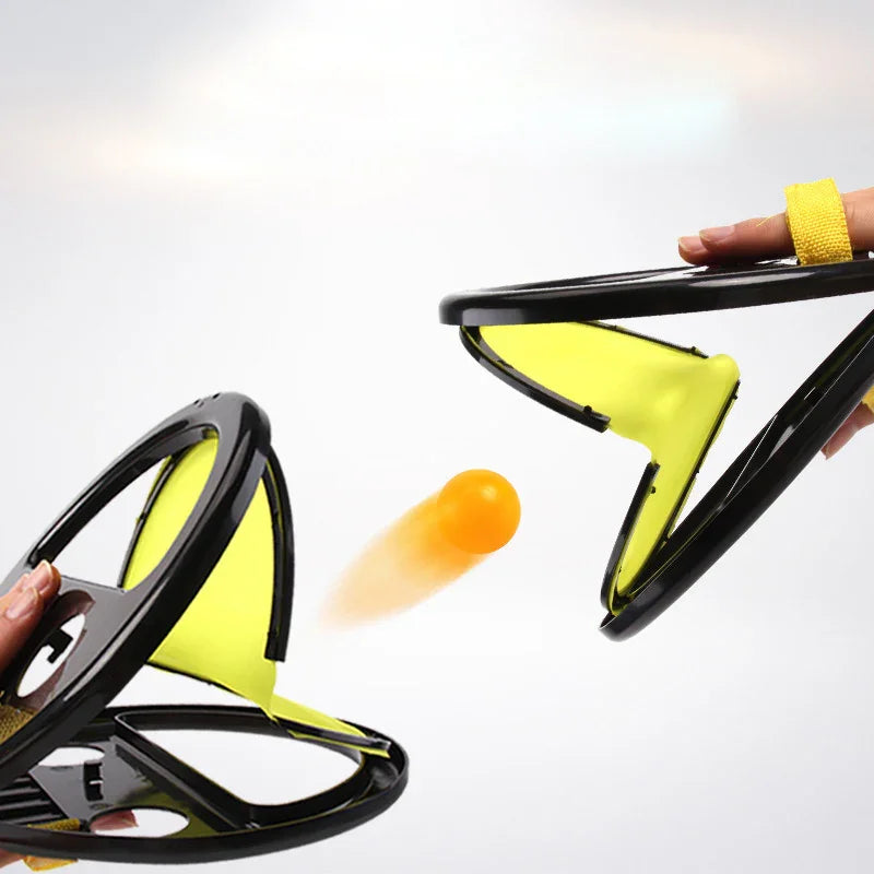 Two hands using yellow and black plastic toy catchers to toss an orange ball, action shot