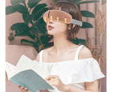 Woman in white off-shoulder top wearing heated eye mask, reading a book indoors