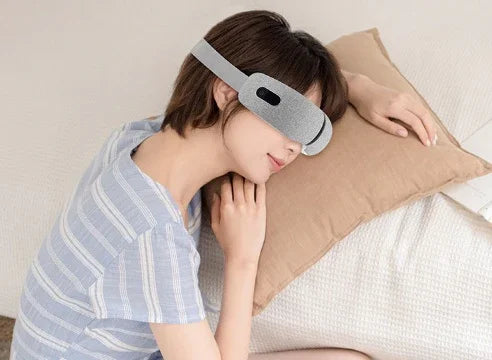 Woman wearing a light gray fabric eye mask with adjustable strap, relaxing in striped loungewear top