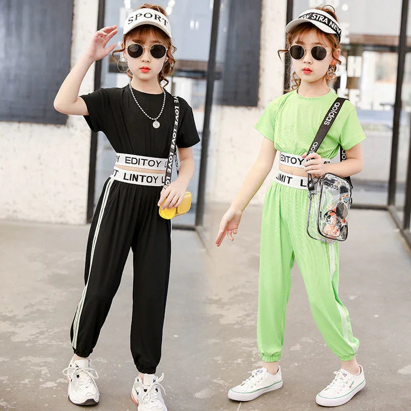 Two girls in sporty streetwear jumpsuits, one black and one neon green, with white stripes, loose fit, and elastic waists, accessorized with visors and sunglasses.