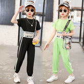 Two girls in sporty streetwear jumpsuits, one black and one neon green, with white stripes, loose fit, and elastic waists, accessorized with visors and sunglasses.