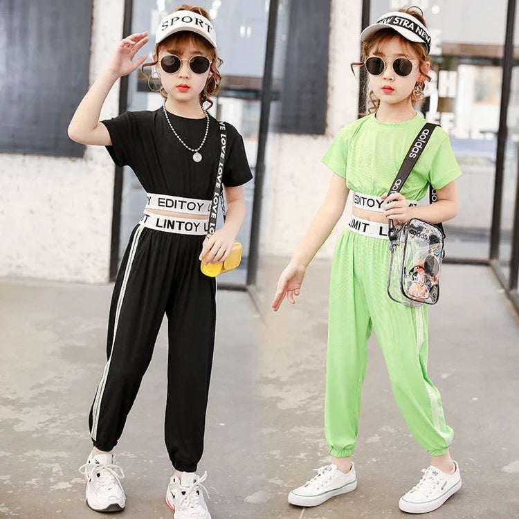 Two girls in sporty streetwear jumpsuits, one black and one neon green, with white stripes, loose fit, and elastic waists, accessorized with visors and sunglasses.