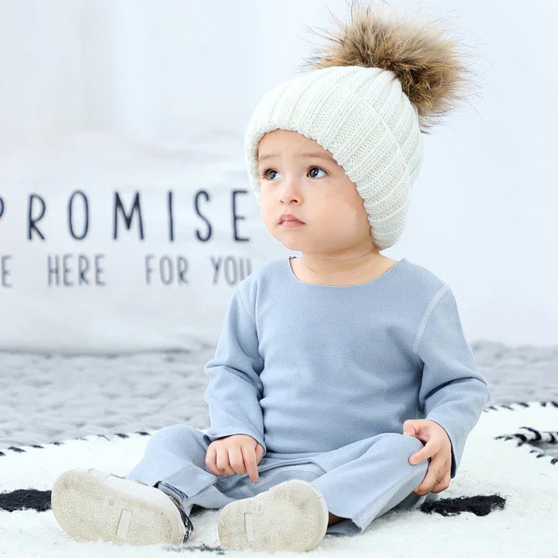 Baby in light blue long sleeve romper and chunky white knit beanie with faux fur pompom
