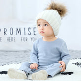 Baby in light blue long sleeve romper and chunky white knit beanie with faux fur pompom