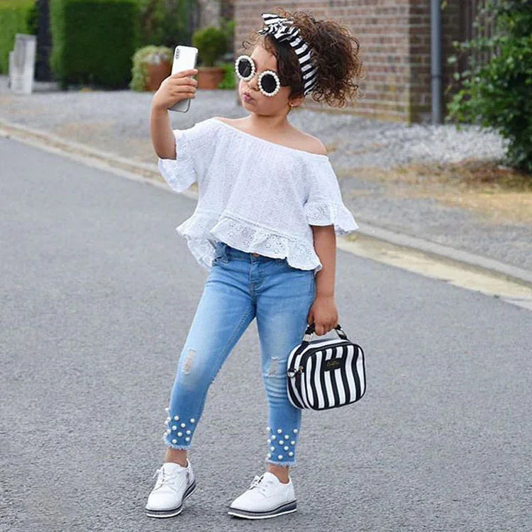 Girl in white off-shoulder lace top, light blue pearl-embellished skinny jeans, white shoes, striped headband and matching handbag, posing outdoors.