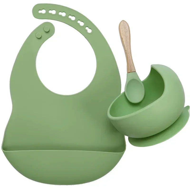 Green silicone baby bib, suction bowl, and spoon set with wooden handle on white background