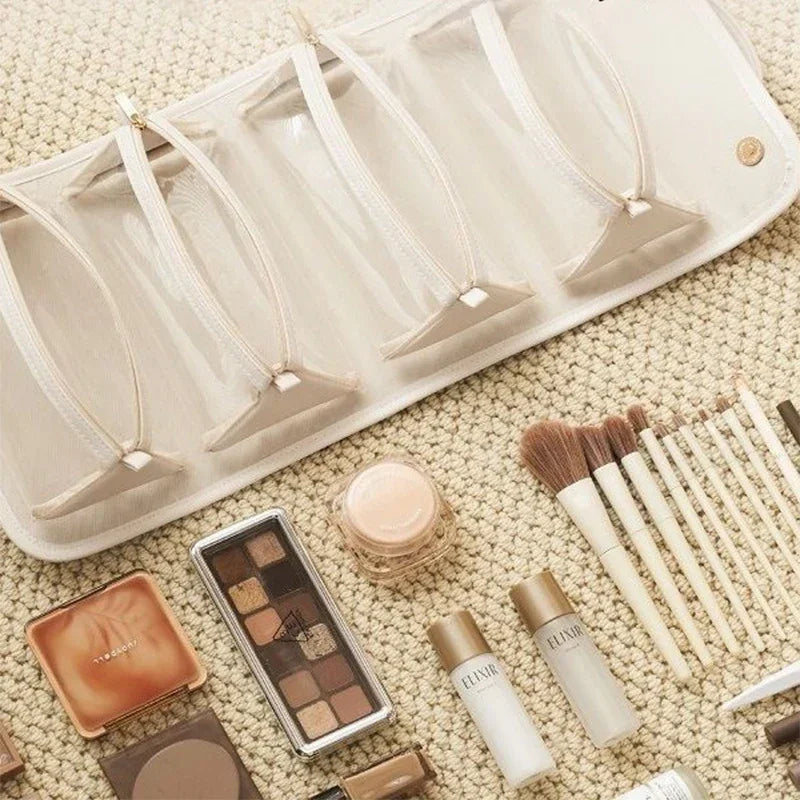 Beige mesh travel makeup organizer with zipper pockets, surrounded by makeup brushes and cosmetics on a textured cream surface.
