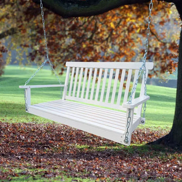 White wooden slat porch swing bench with chains hanging from a tree in a park setting.