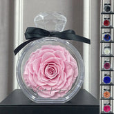 Decorative pink preserved rose in clear round case with black ribbon and diamond-shaped lid.