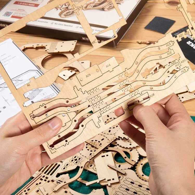 Hands holding laser-cut wooden model kit pieces on a desk with assembly parts and instructions