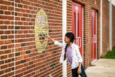 Girl in casual jeans and light jacket with purple shirt, drawing on brick wall with chalk art.