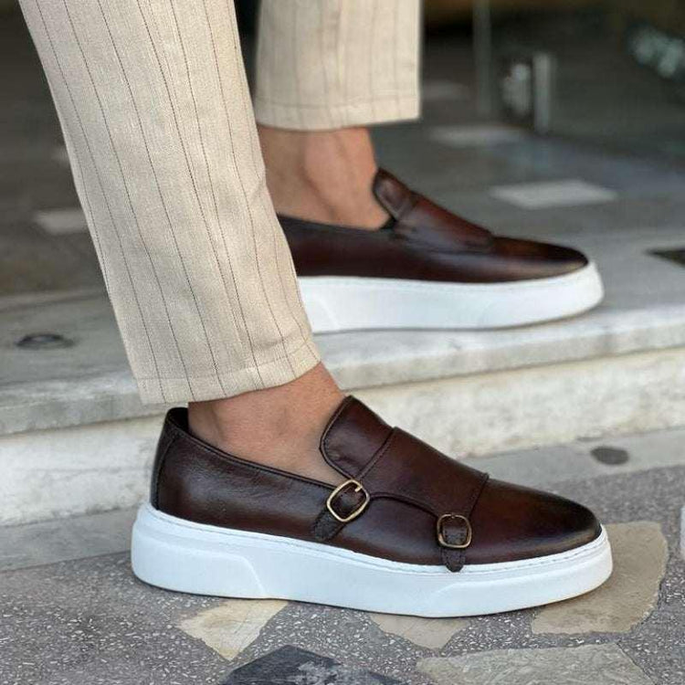 Brown leather slip-on sneakers with white soles and gold buckle details, worn with beige striped pants