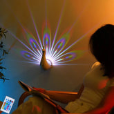 Woman in a fitted white short-sleeve top reading by a peacock-shaped LED wall light projecting colorful patterns.