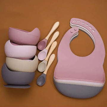 Stacked silicone baby bowls, matching spoons with wooden handles, and silicone bibs in muted pastel colors.