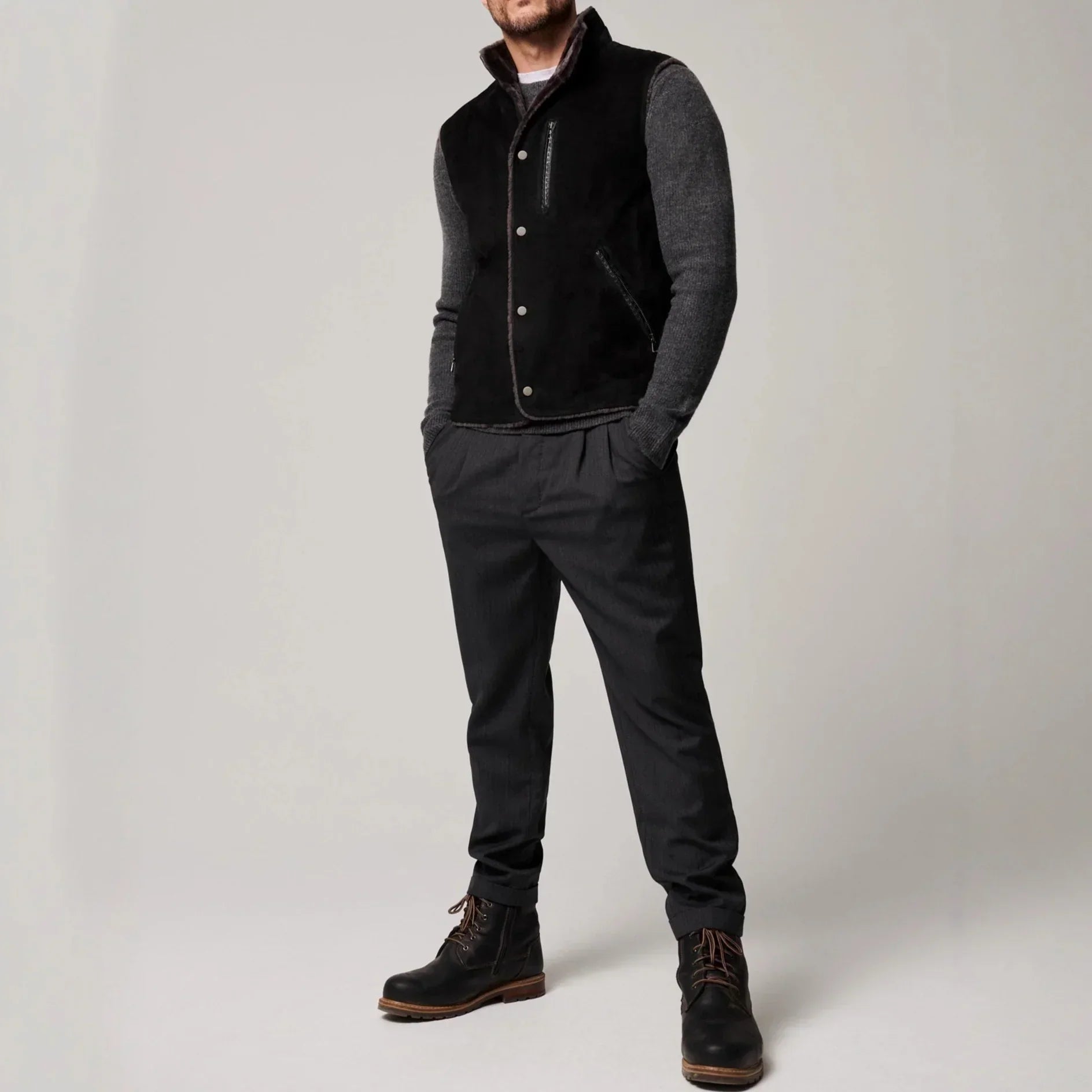 Man wearing a black suede vest, grey knit sleeves, dark tapered pants, and black boots.