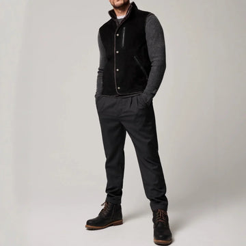 Man wearing a black suede vest, grey knit sleeves, dark tapered pants, and black boots.