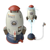 Rocket-shaped water sprinkler toy in white and red plastic, connected to a garden hose, spraying water upwards.