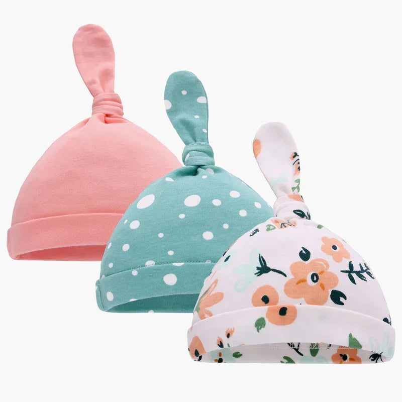 Three soft cotton baby hats with knotted tops, in pink, green polka dot, and floral prints.