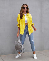 Woman in oversized yellow distressed denim jacket, white crop top, blue ripped skinny jeans, and white sneakers, holding a gray backpack.